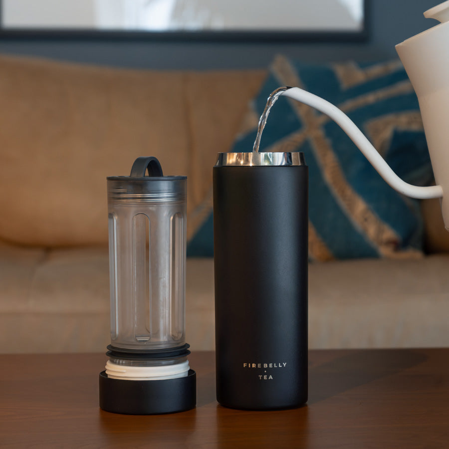 Pouring hot water into a black 'Firebelly Tea' infuser bottle on a table.